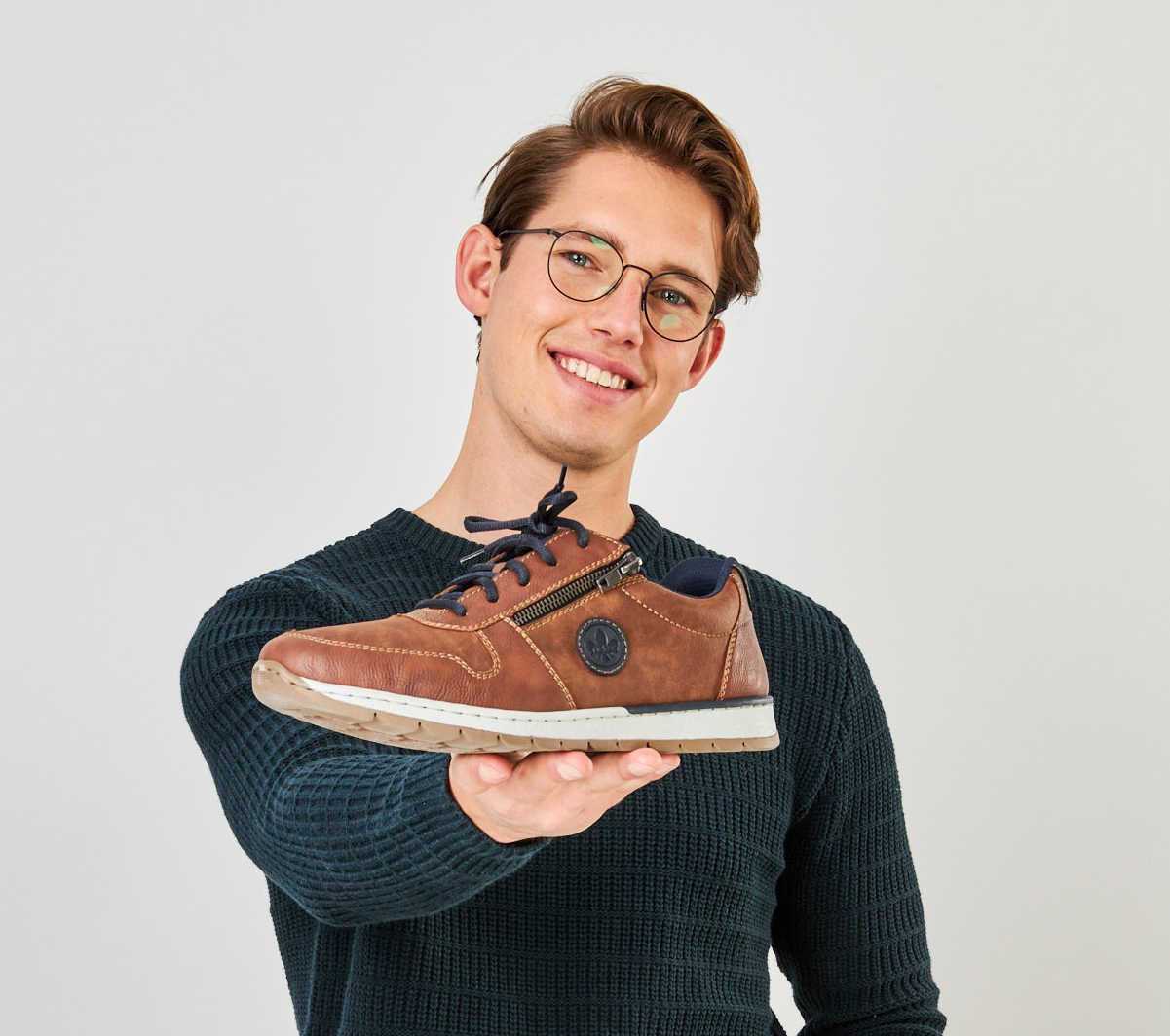 Ein lächelnder junger Mann hält einen braunen Schuh ins Bild