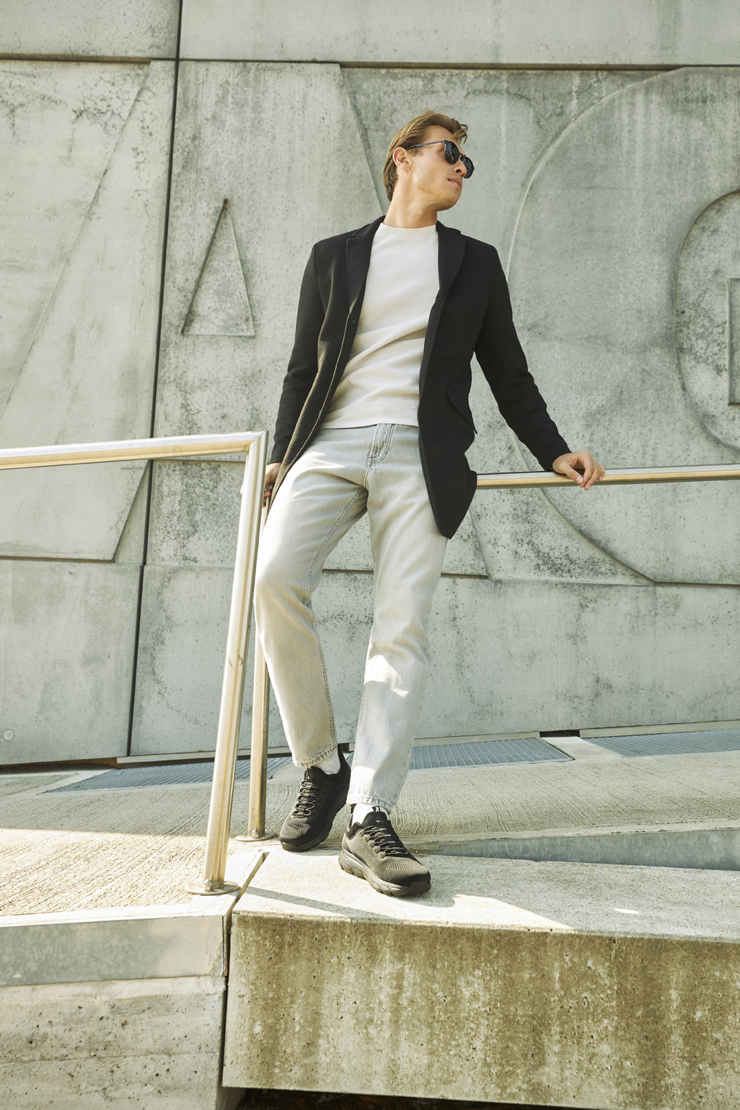 Schwarze R-EVOLUTION Slipper von Rieker aus Textil mit Gummizug, extra softer und herausnehmbarer Einlegesohle sowie super leichter und flexibler Laufsohle. Passend zu den Schuhen trägt der blonde Mann eine helle Jeans mit einem weißen Pullover und einem schwarzen Mantel. Er steht an einem Geländer.