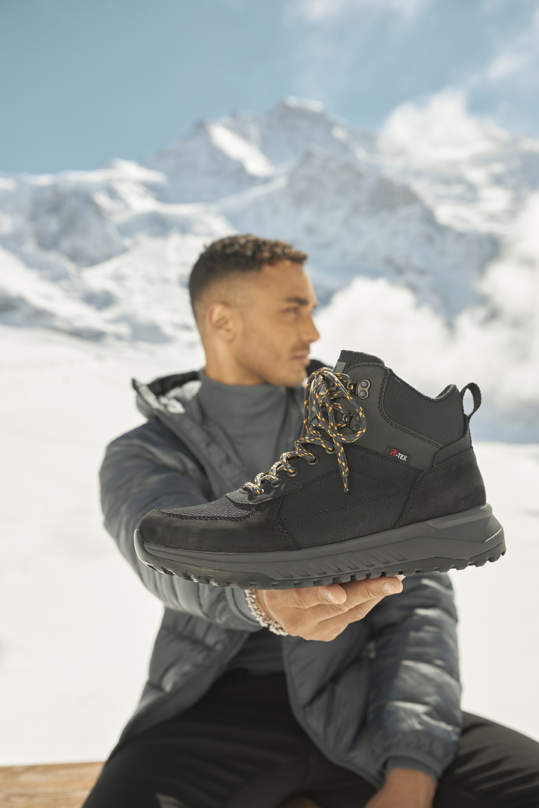 [Translate to Englisch (USA):] Black R-EVOLUTION boots by Rieker made of synthetic leather with zipper, extra soft and removable insole and super light and flexible outsole. Matching the shoes, the dark-haired man is wearing black jeans with a grey turtleneck sweater, sunglasses and a grey vest while sitting on a bench in the snow.